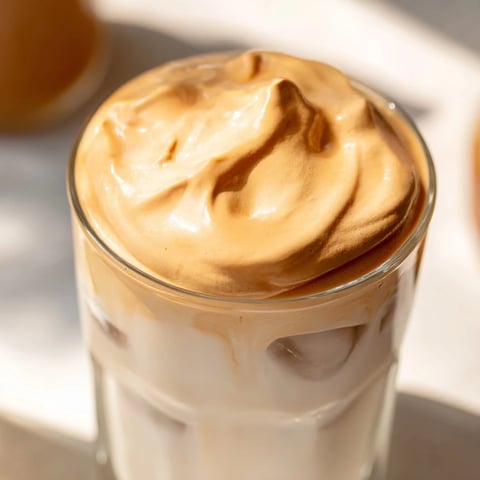 Creamy whipped Dalgona coffee floats atop cold milk in a glass, topped with ice cubes for a refreshing café-style treat.  